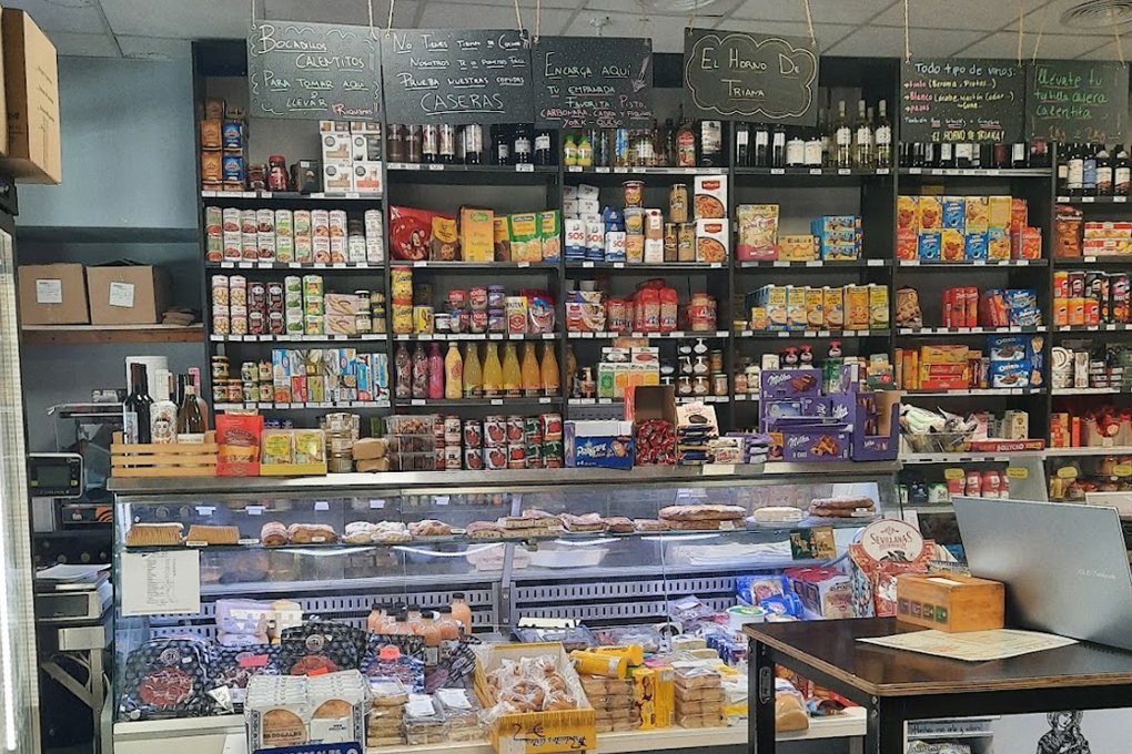 Interior de la tienda con estanterías de alimentación, vitrina de dulces y pizarras escritas a tiza, una con el nombre El Horno de Triana